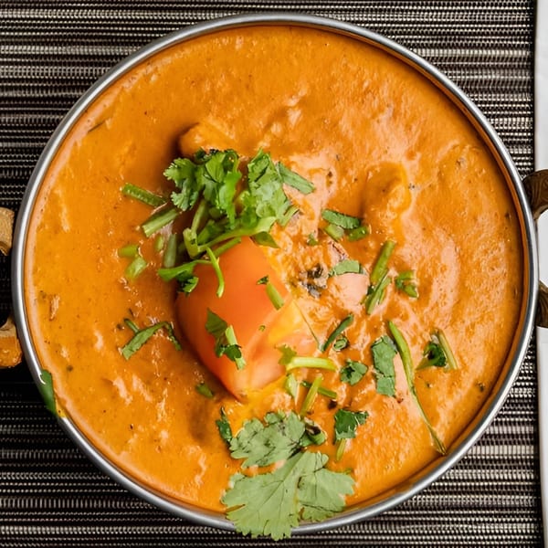Overhead view of a vibrant orange curry in a metal bowl, garnished with cilantro and a tomato wedge.